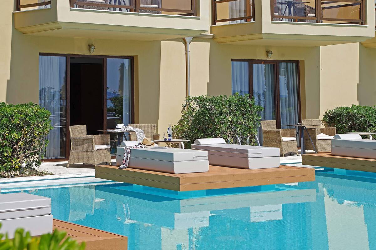 A poolside area with lounge chairs and a small table, surrounded by greenery and a building with balconies.