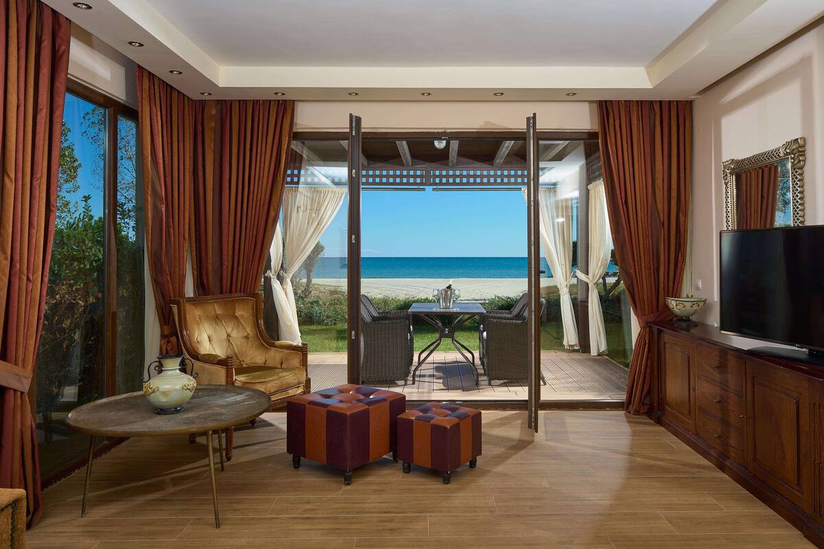 A room with a view of the beach, a TV, a table, and two ottomans.