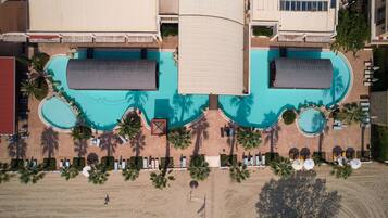 An aerial view of a resort with a large central pool, multiple smaller pools, and a beach area with sun loungers and umbrellas.