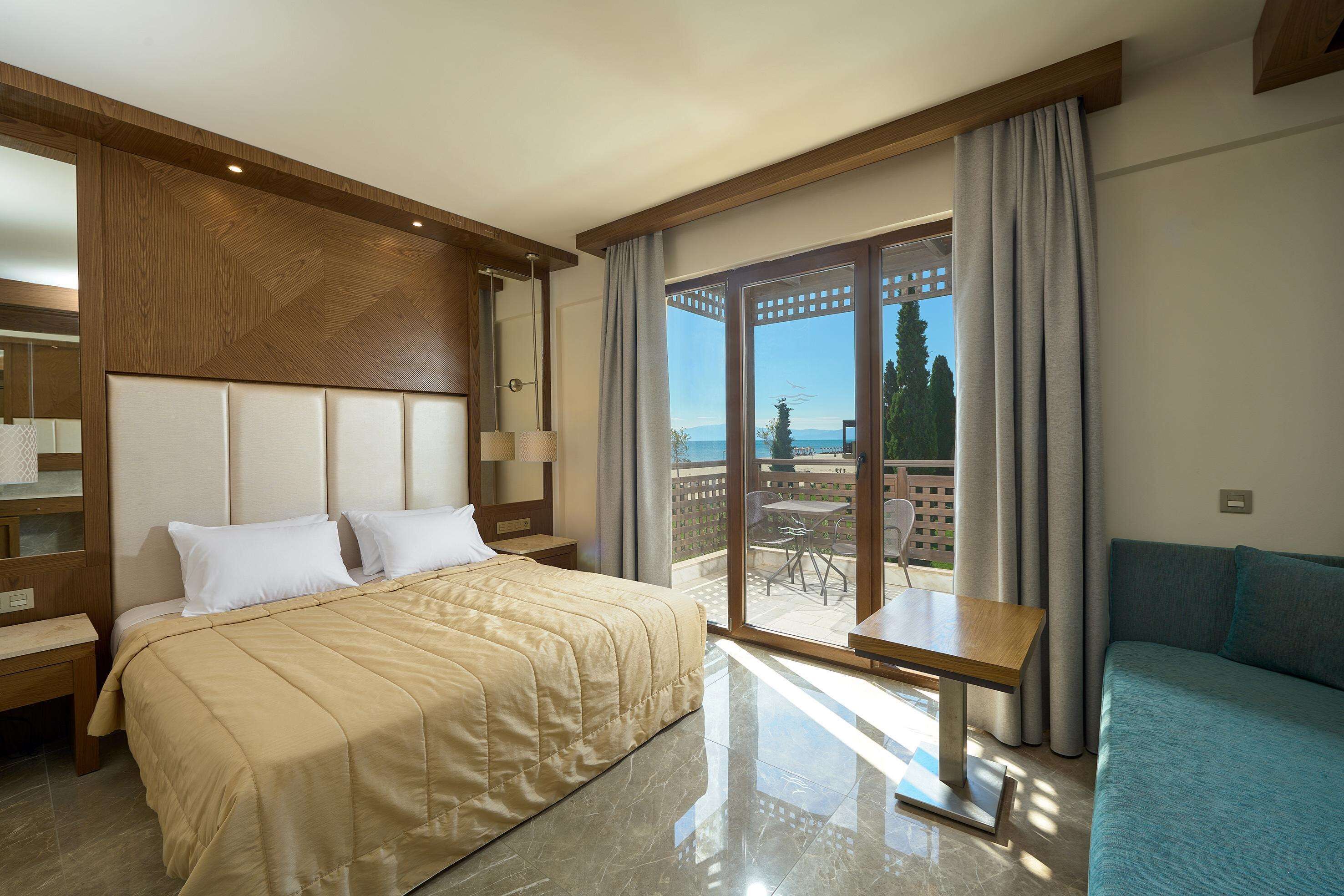 A hotel room with a bed, a sofa, a balcony with a table and chairs, and a view of the outdoors.