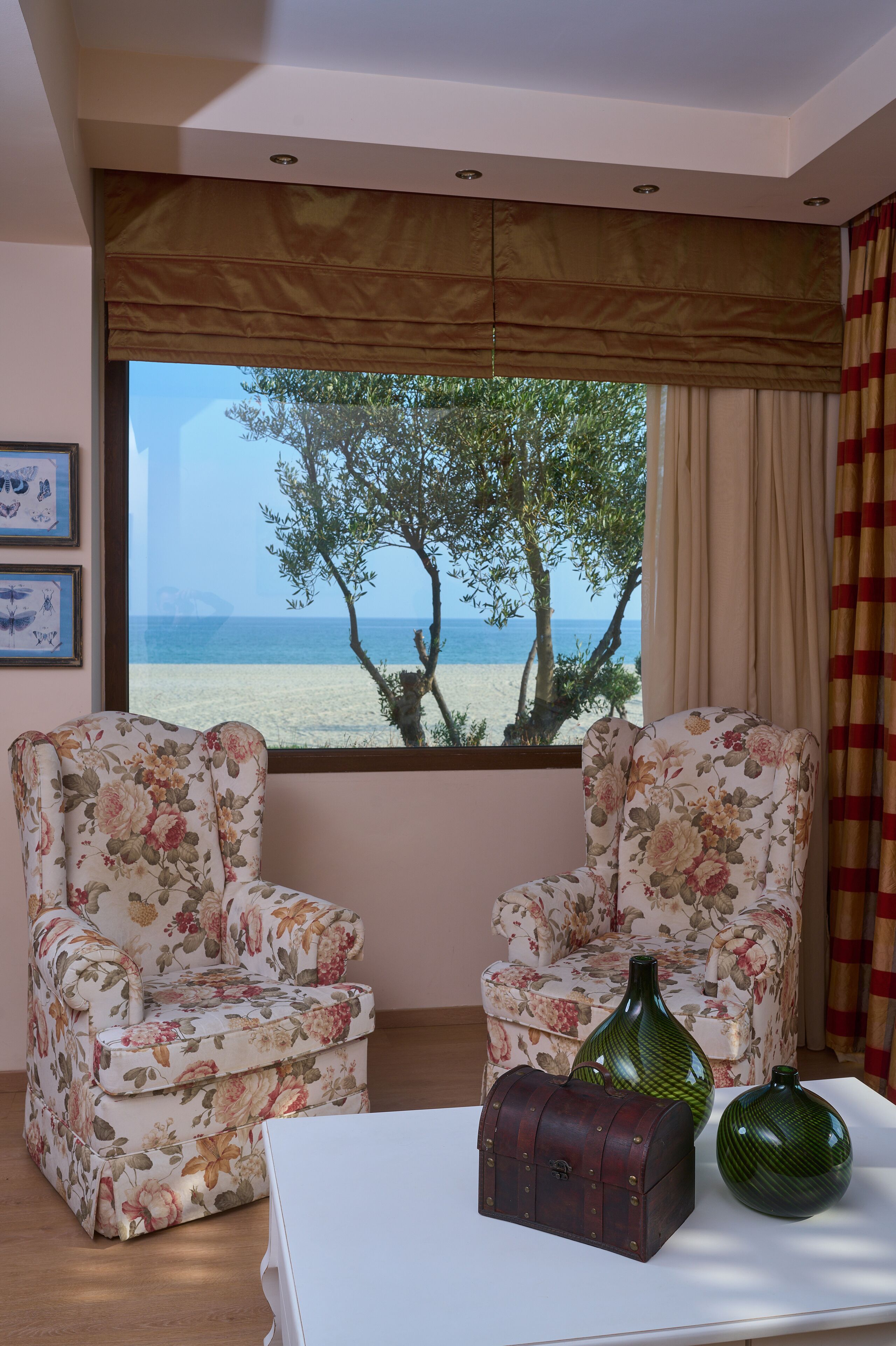 A room with two floral armchairs, a white table, and a view of the beach and sea through a large window.