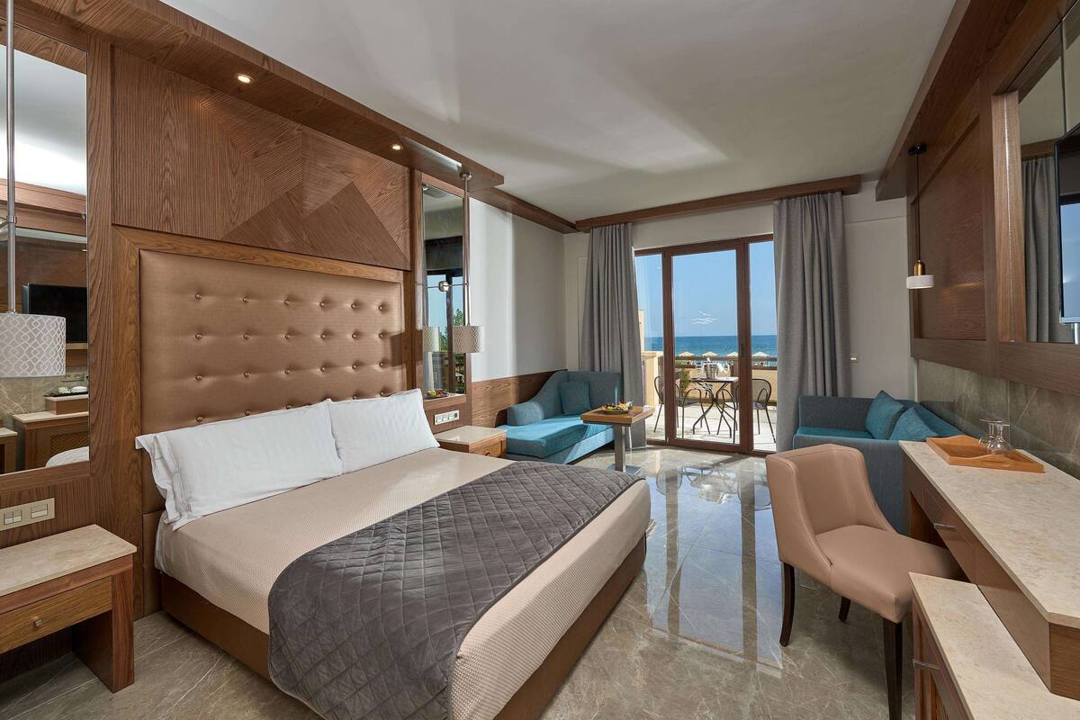 A hotel room with a large bed, a desk, a chair, and a balcony with a view of the sea.