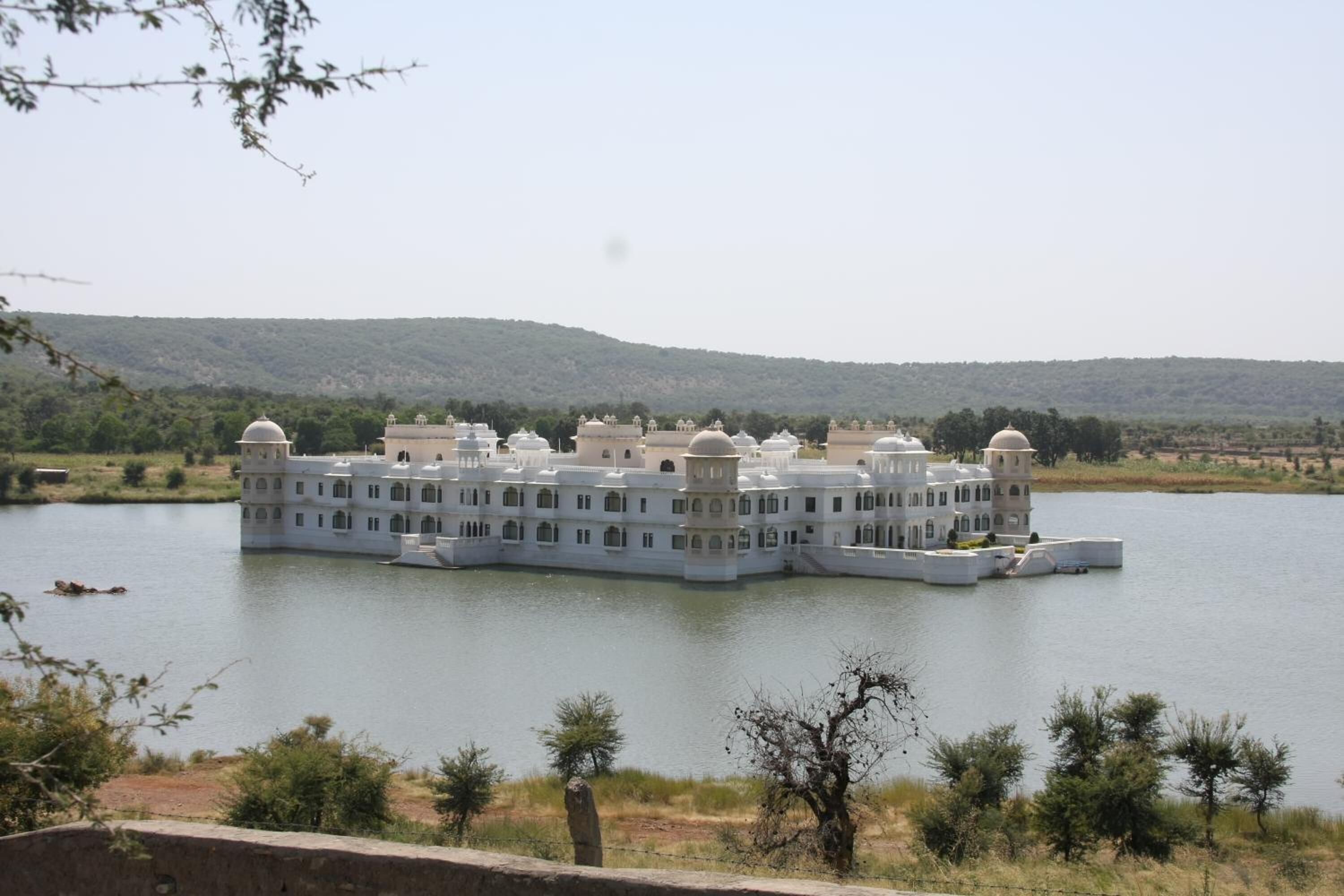 A large, white, multi-story building with domed roofs, situated on a lake with a hilly backdrop.