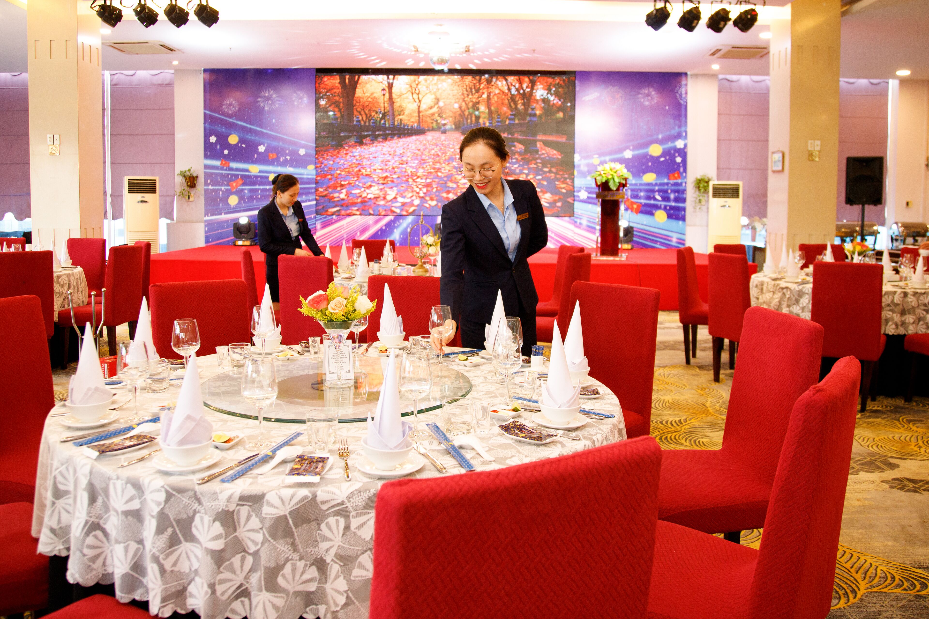 Un salón de banquetes con mesas redondas dispuestas para comer, sillas tapizadas en rojo y una pantalla grande mostrando un paisaje.