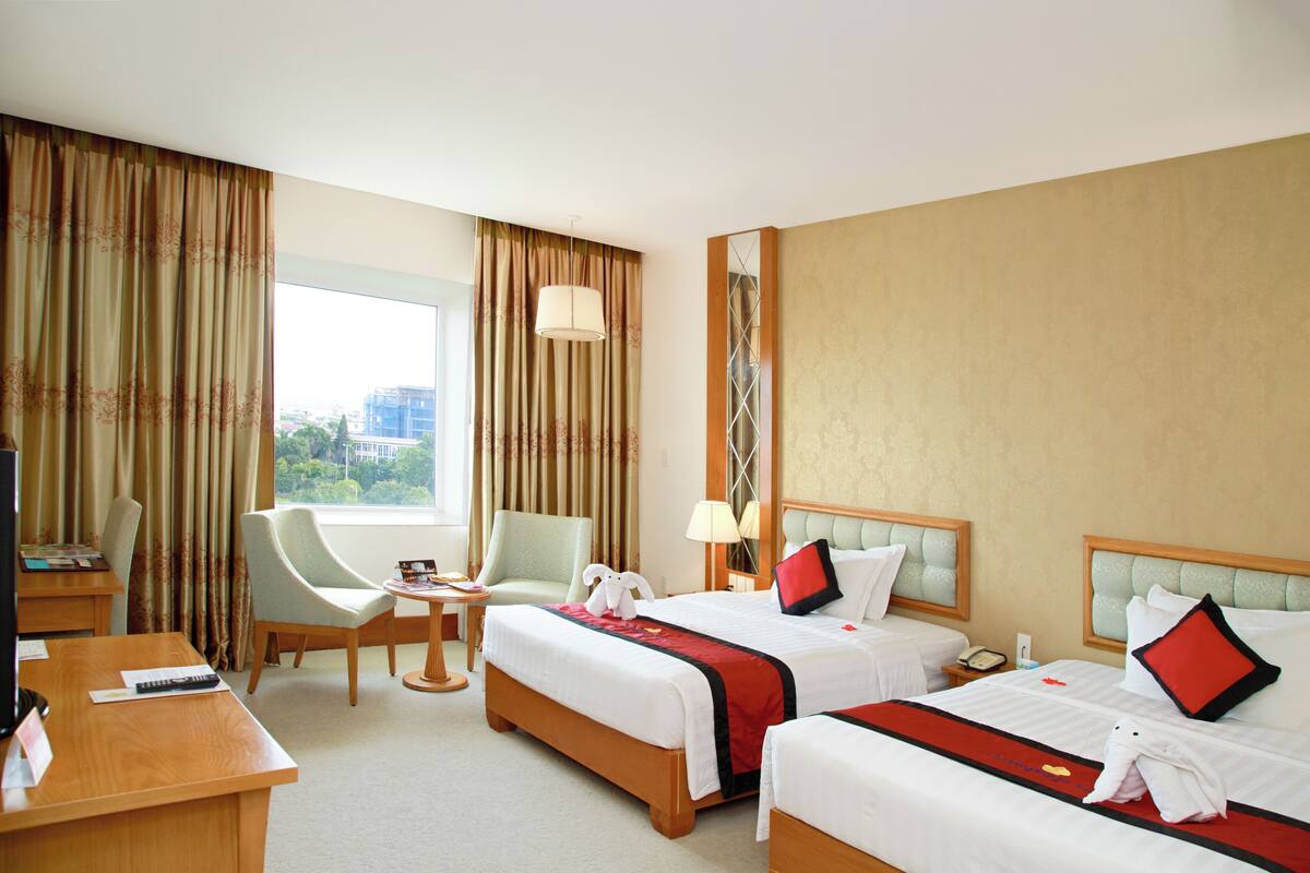 A hotel room with two beds, a desk, a chair, a lamp, and a view of the city.