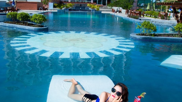 Una persona descansando en una silla de piscina junto a una alberca, con un edificio de hotel al fondo.