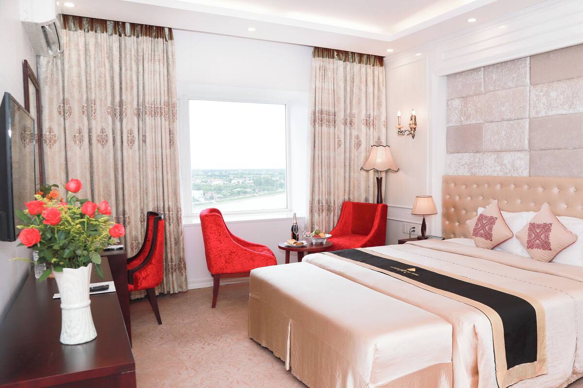 A hotel room with a large bed, a red chair, a desk with a vase of flowers, and a view of the city through the window.