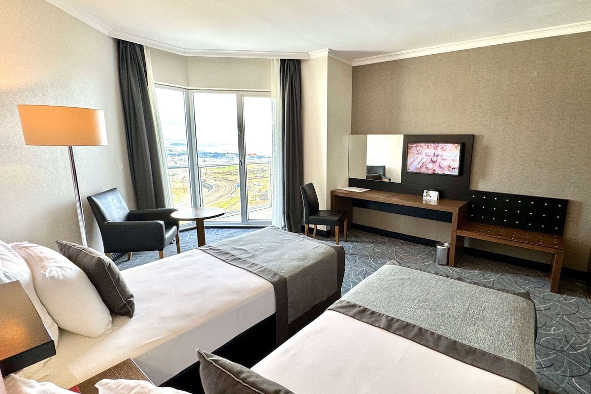A hotel room with two beds, a desk, a chair, a TV, and a view of the city through the window.
