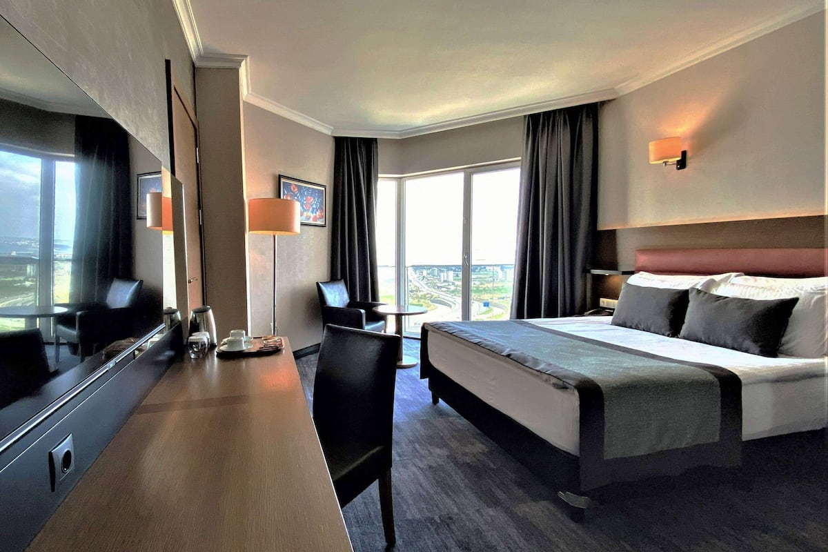 A hotel room with a large bed, a desk with a chair, a coffee maker, and a large window with curtains.