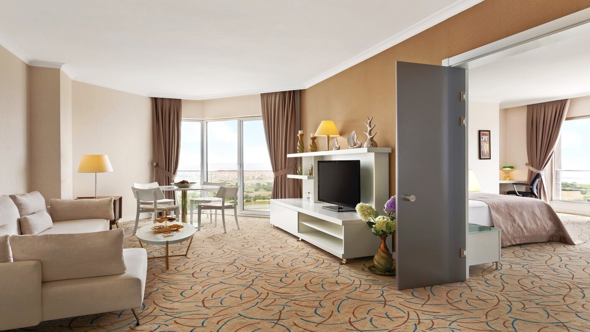 Habitación de hotel con sofá, mesa de comedor con sillas, televisor en un mueble bajo y cama con vista a la ciudad.