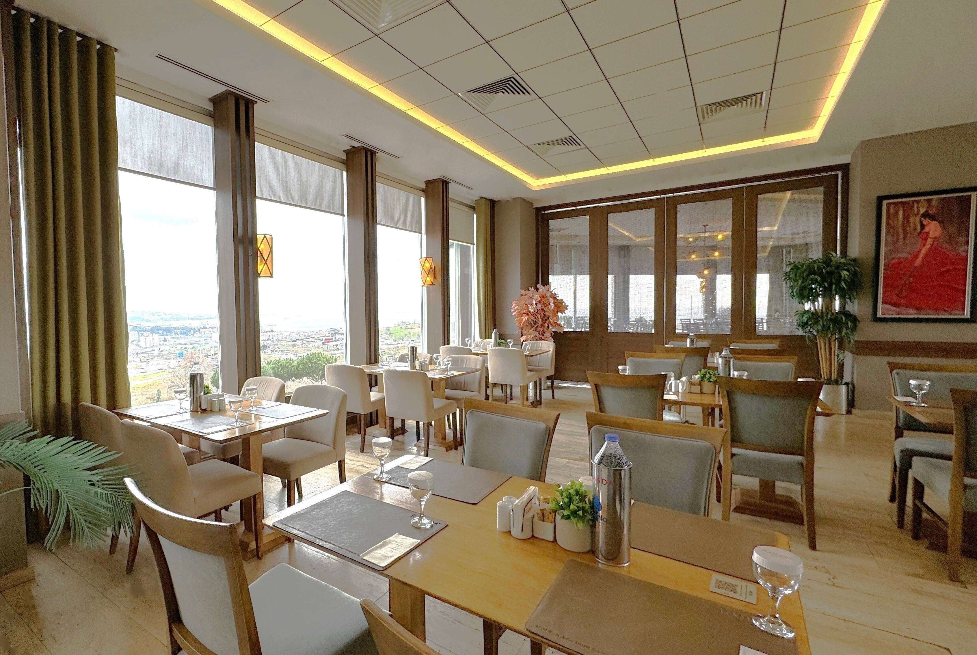 A hotel dining area with multiple tables and chairs, large windows offering a city view, and a modern ceiling design.
