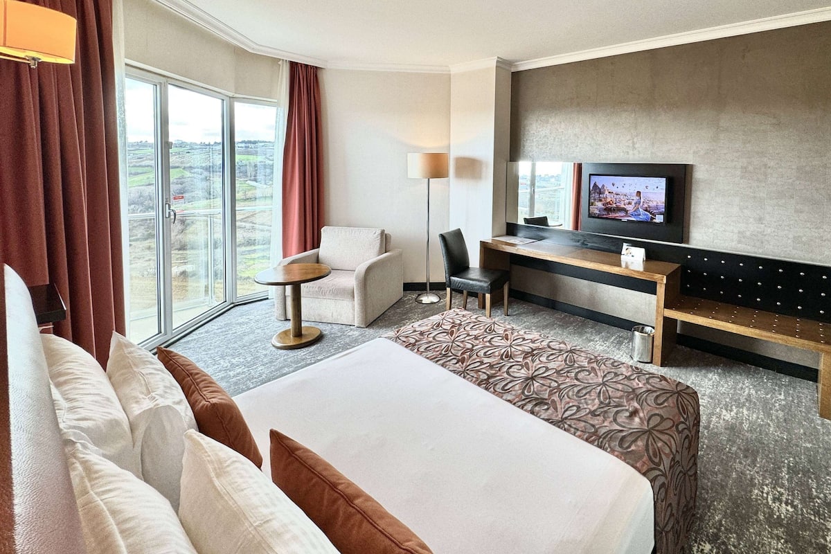 A hotel room with a large bed, a desk with a television, a chair, and a view of the city through large windows.