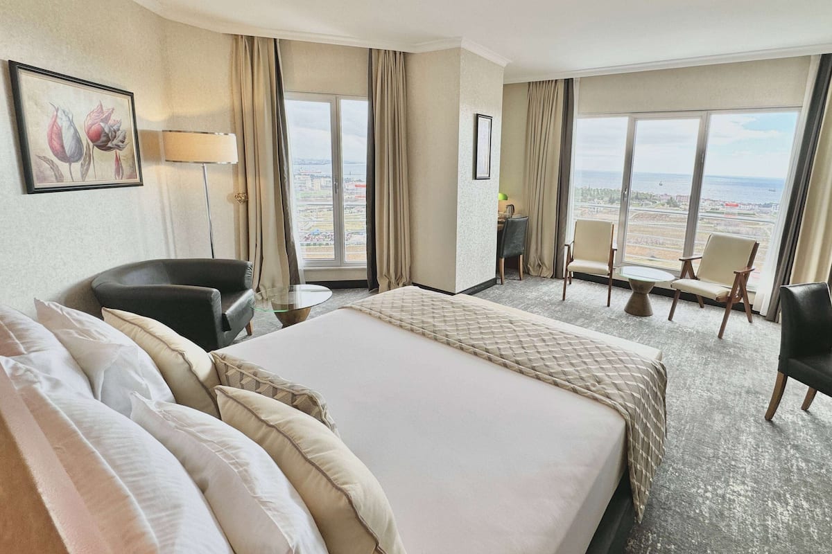 A hotel room with a large bed, a desk, a chair, and a view of the city through large windows.