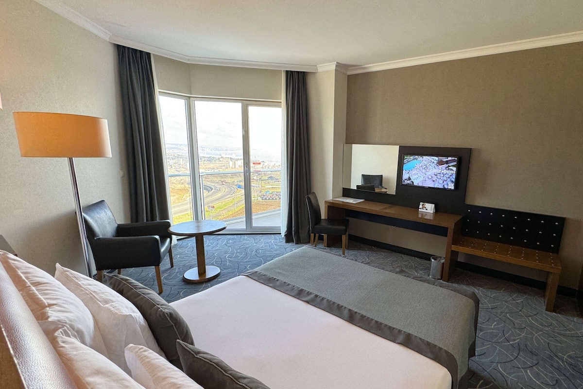 A hotel room with a large bed, a desk with a television, a chair, and a view of the city through the window.