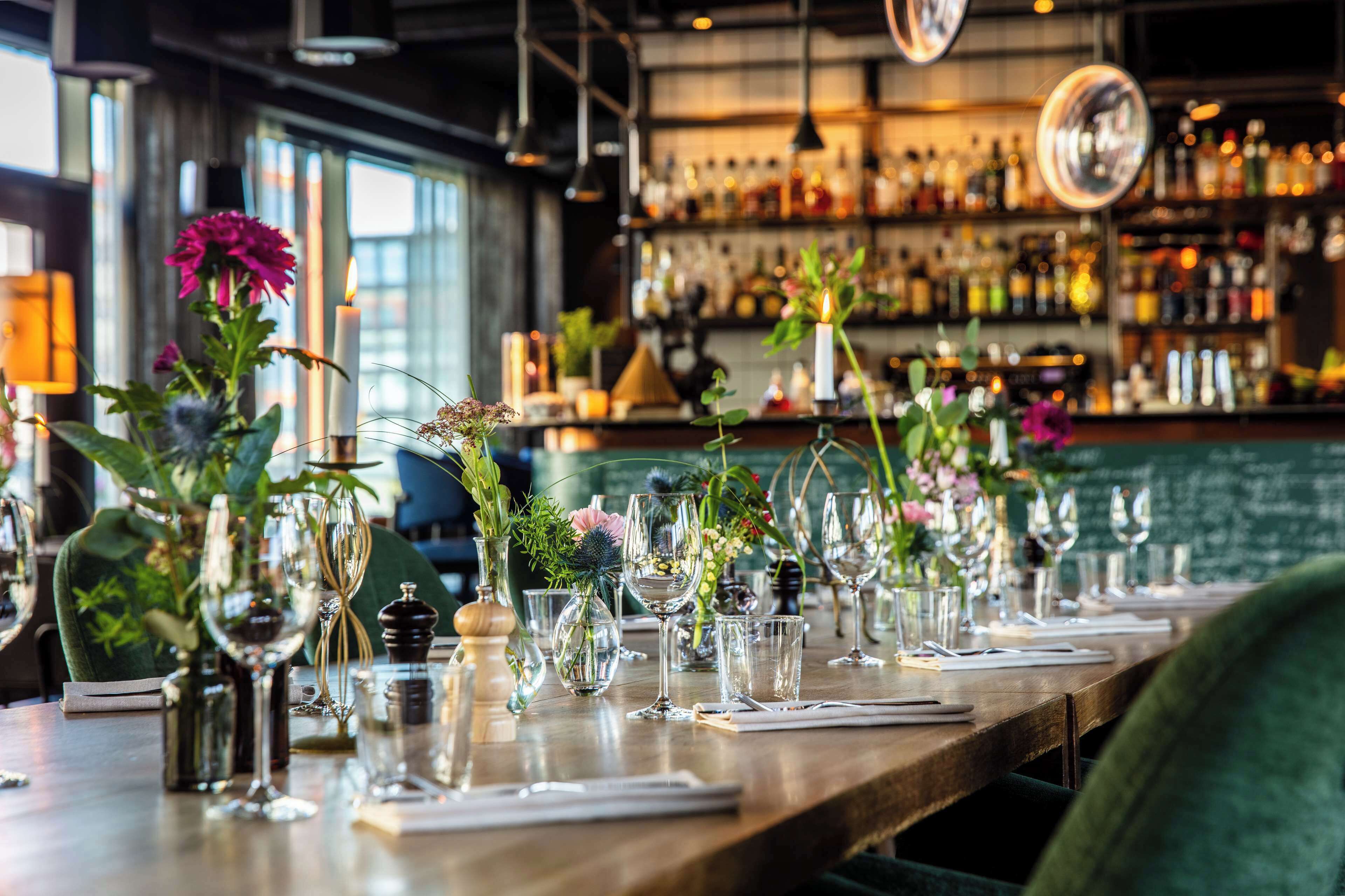 A bar setting with a long wooden counter, multiple wine glasses, and various decorative vases with flowers.