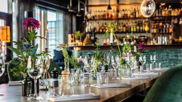 A bar setting with a long wooden counter, multiple wine glasses, and various decorative vases with flowers.