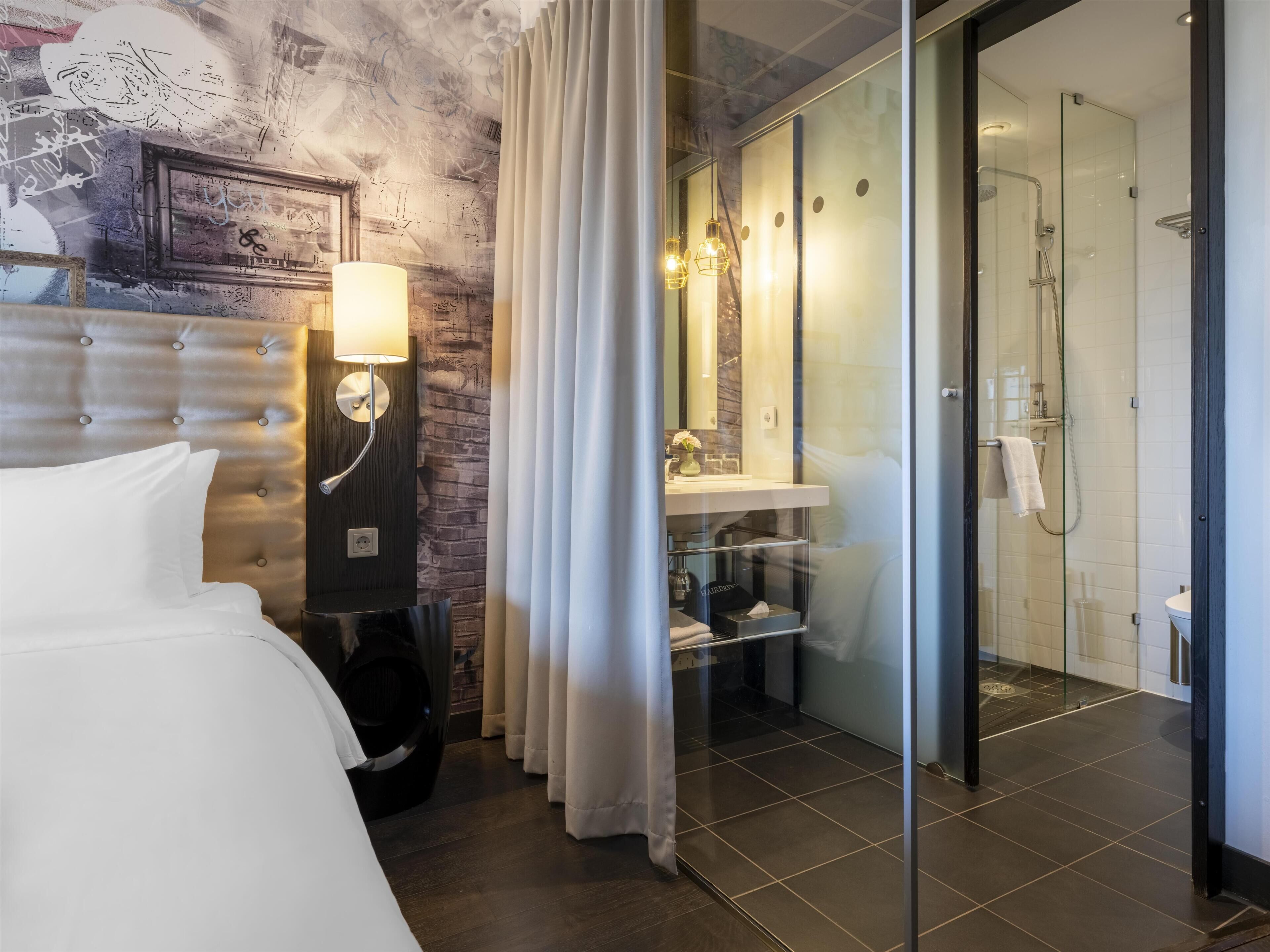 A hotel room with a bed, a wall-mounted lamp, a bathroom with a glass shower enclosure, and a visible door.