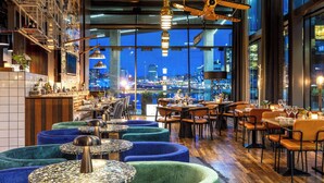 En restaurang med runda bord, blå och gröna, vadderade sittplatser och utsikt över staden genom stora fönster.