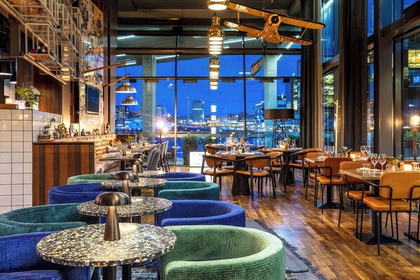 En restaurang med runda bord, blå och gröna, vadderade sittplatser och utsikt över staden genom stora fönster.