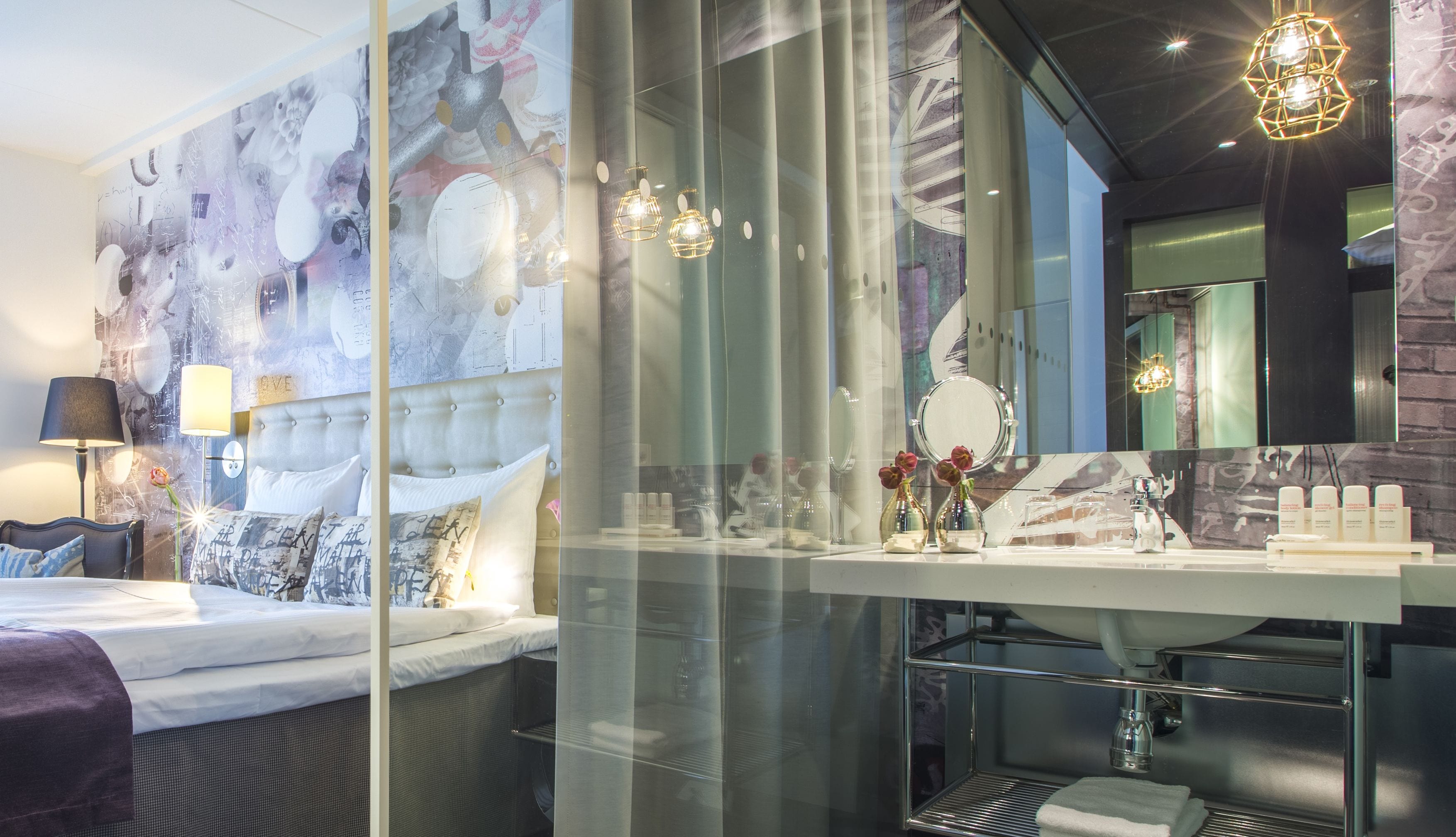 A modern hotel room with a large bed, a glass shower enclosure, a vanity with a mirror, and decorative items.