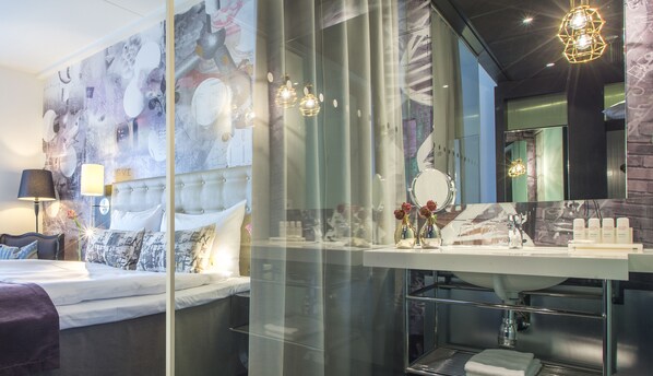 Una habitación de hotel moderna con una cama grande, cabina de ducha de vidrio, tocador con espejo y elementos decorativos.