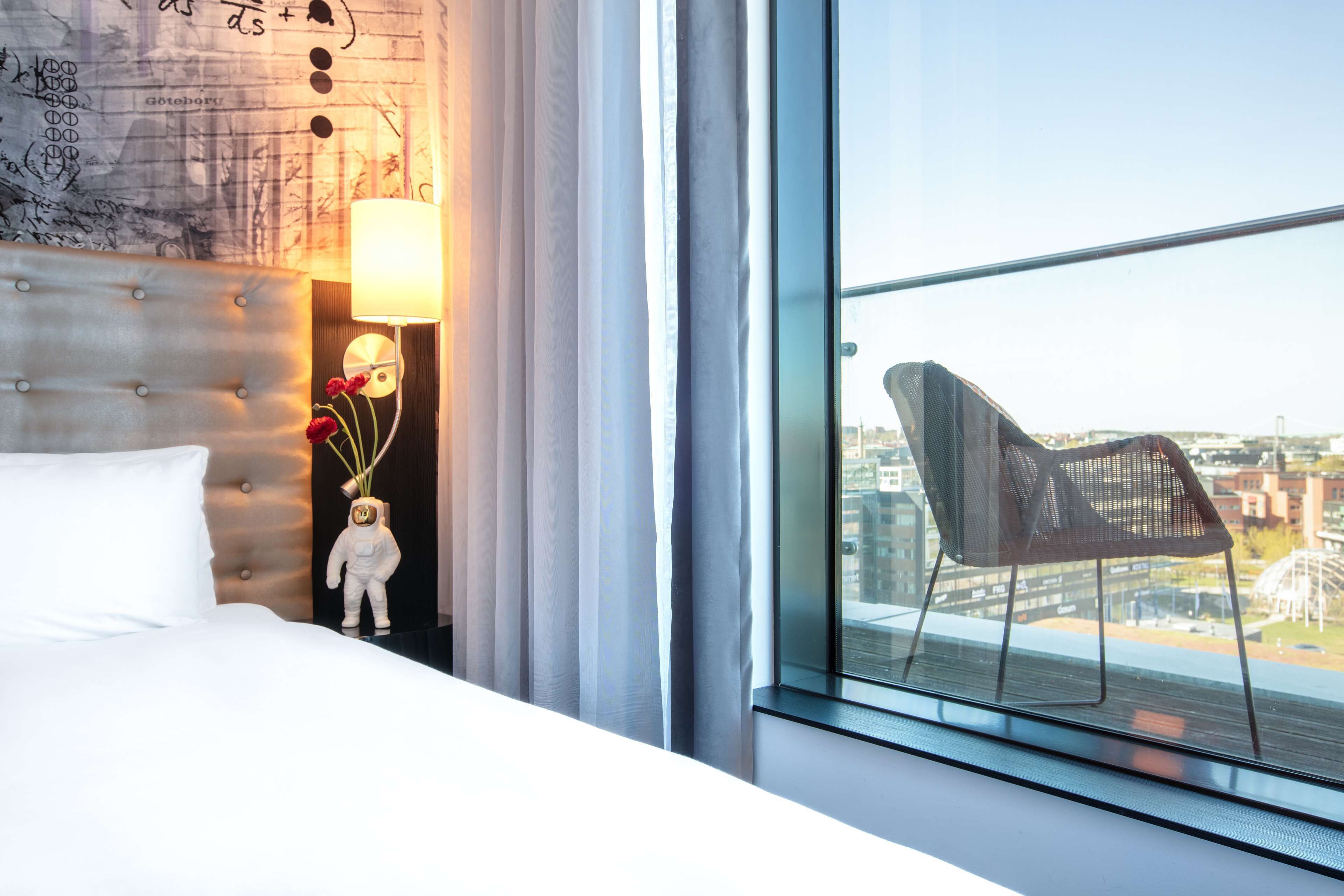 Hotelkamer met een groot raam, een bed met witte lakens, een wandlamp en een decoratief object op het nachtkastje.
