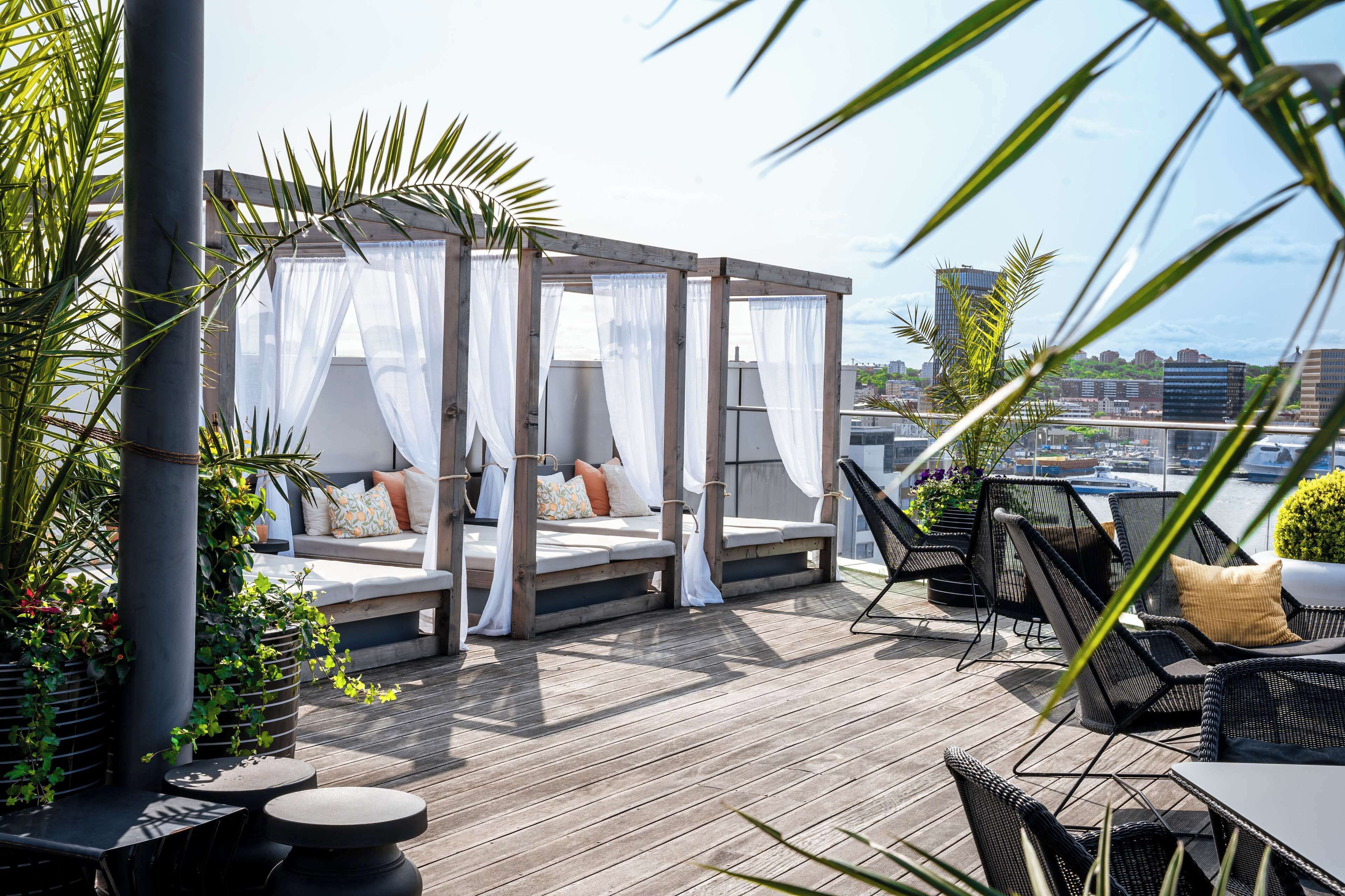 A rooftop terrace with a wooden deck, outdoor seating with cushions, and a view of the city skyline.