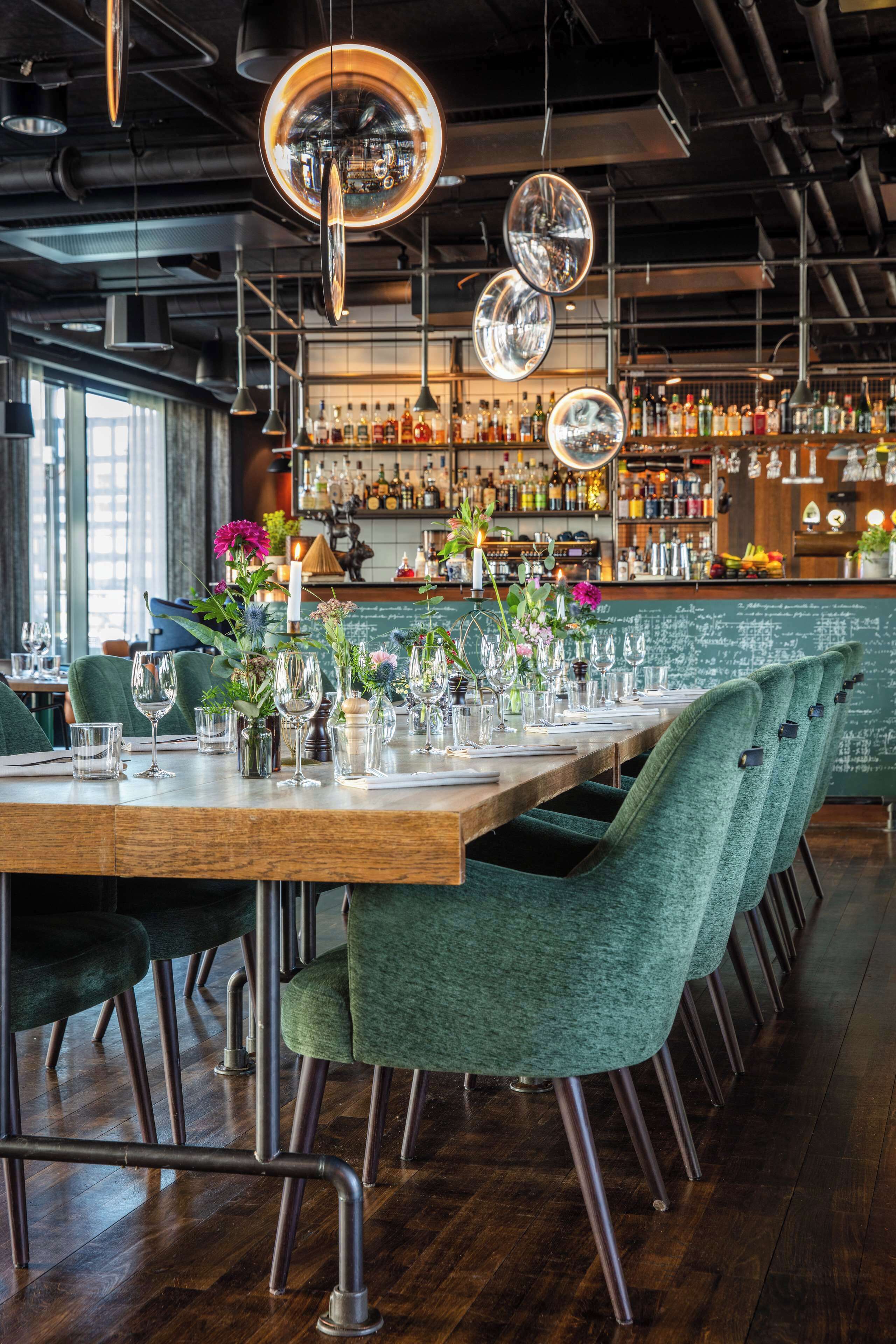 En restaurant med et langt træbord dækket til spisning, grønne polstrede stole og et barområde med flasker på hylder.