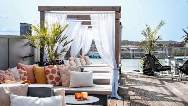 A rooftop terrace with a wooden deck, a seating area with cushions and a table, and a view of the marina and buildings in the background. - Radisson Blu Riverside Hotel, Gothenburg (Gothenburg)
