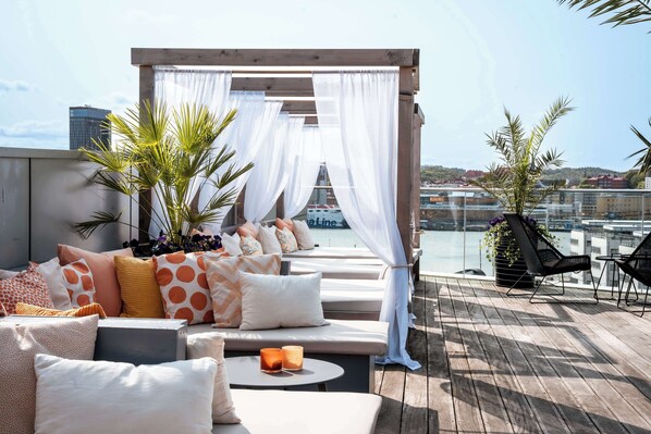 A rooftop terrace with a wooden deck, a seating area with cushions and a table, and a view of the marina and buildings in the background. - Radisson Blu Riverside Hotel, Gothenburg (Gothenburg)