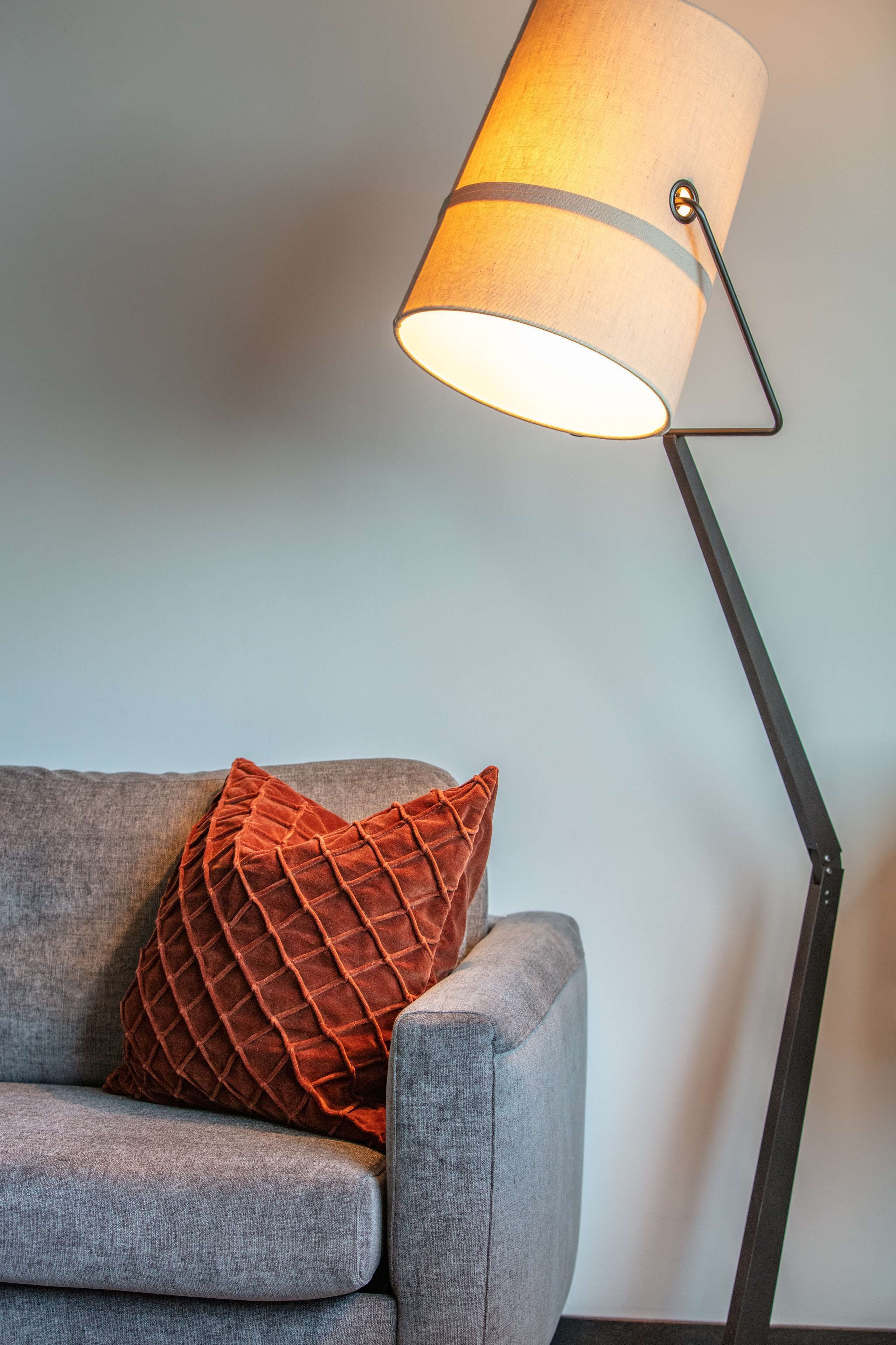 A modern lamp with a cylindrical shade and a black stand, positioned next to a grey sofa with a textured orange pillow.