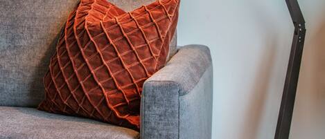 A modern lamp with a cylindrical shade and a black stand, positioned next to a grey sofa with a textured orange pillow.