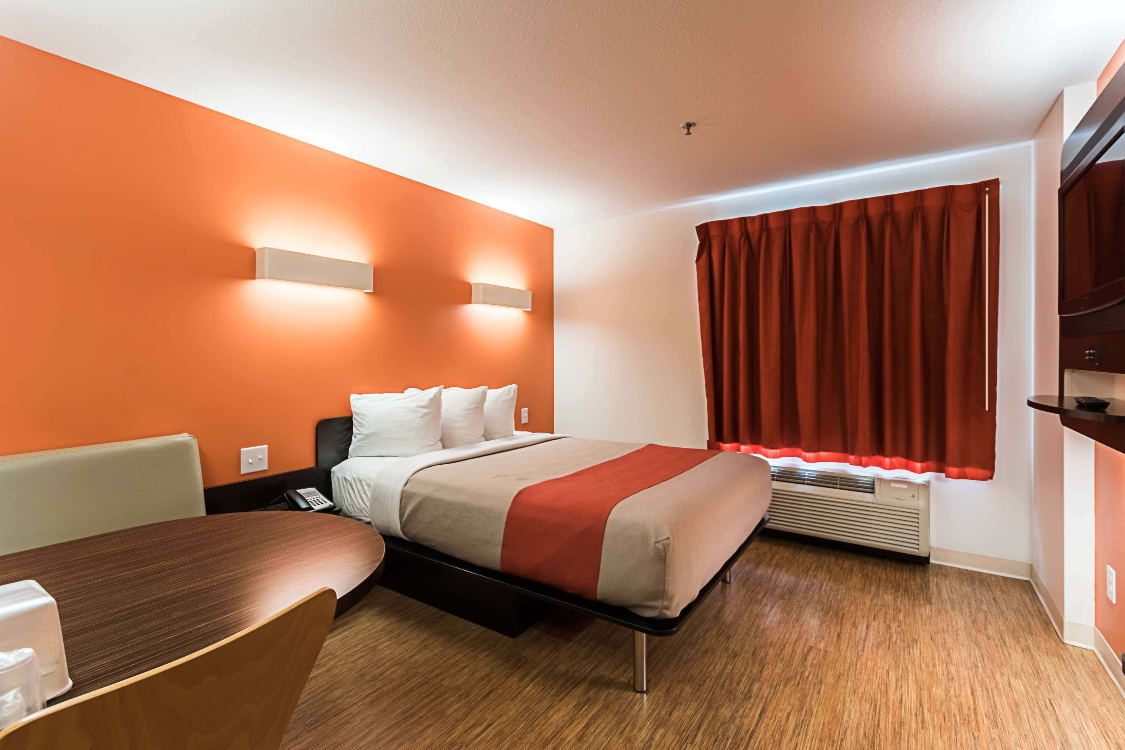 Quarto de hotel com uma cama, uma escrivaninha e uma cadeira. Há uma janela com cortinas e um ar condicionado fixado na parede.