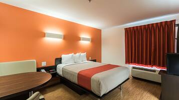 A hotel room with a bed, a desk, a chair, a heater, and a window with curtains.