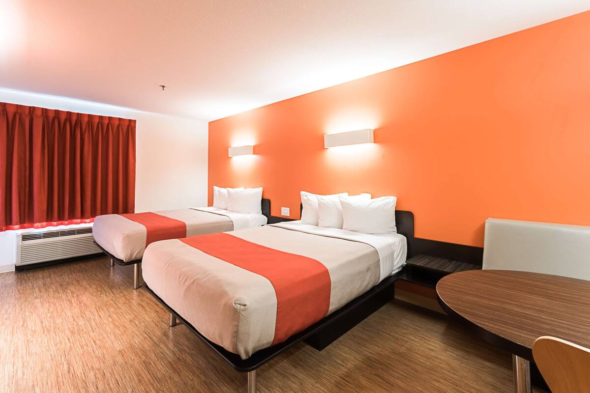 A hotel room with two beds, a wooden floor, an orange accent wall, and a dining table.