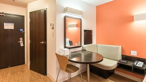 Habitación de hotel con puerta de madera, una mesa redonda con una silla, una pequeña kitchenette y un área de descanso con un banco y un teléfono.