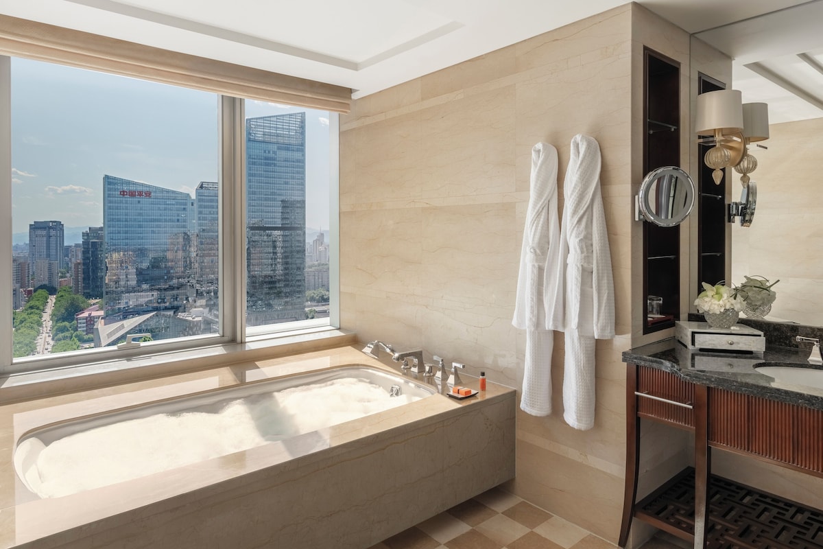 A modern bathroom with a large bathtub, a vanity with a mirror, and a view of a cityscape through a large window.
