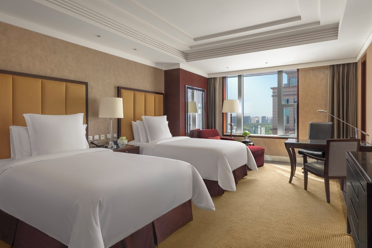 A hotel room with two beds, a desk, a chair, a TV, and a large window with a city view.