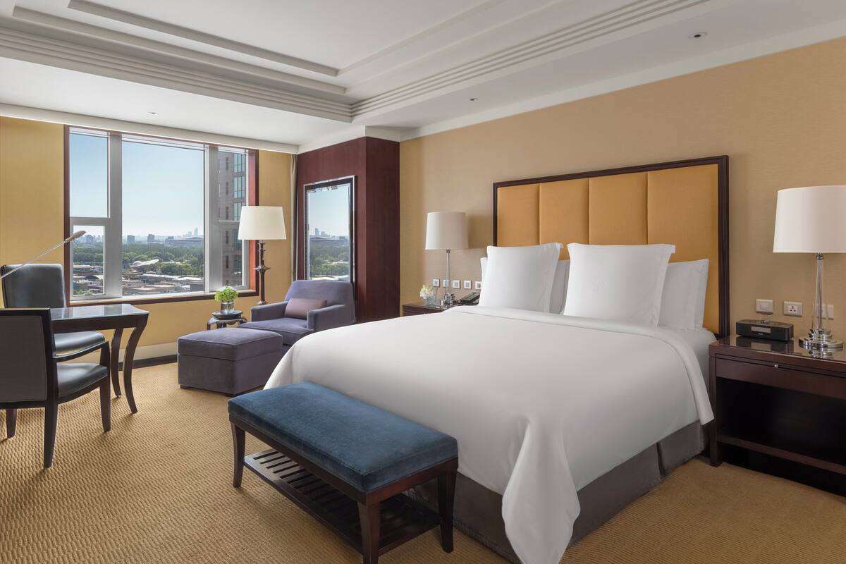 A hotel room with a large bed, a desk, a chair, a bench, and a view of the city through large windows.