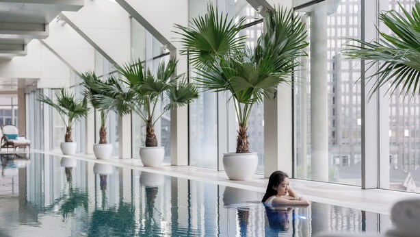 An indoor swimming pool with a view of a cityscape through large windows, surrounded by potted palm plants. - Four Seasons Hotel Beijing (Beijing)
