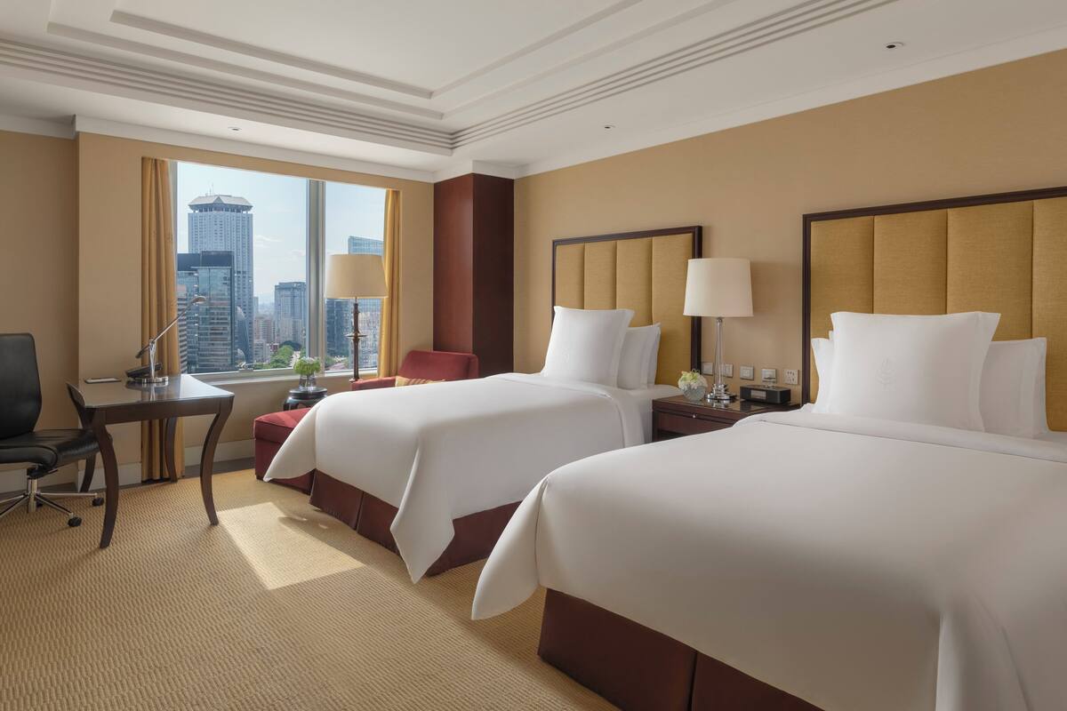 A hotel room with two beds, a desk, a chair, a lamp, and a view of a cityscape.