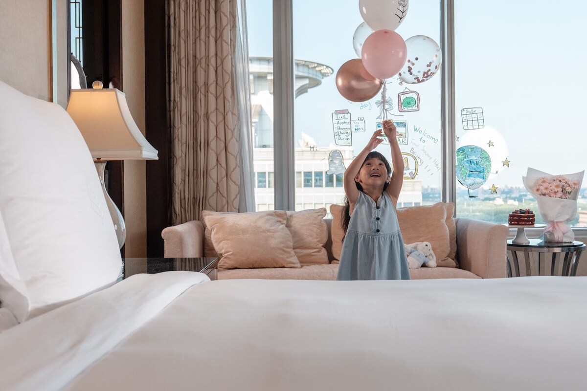 A hotel room with a bed, a sofa, a lamp, a window with a view of a building, and a wall decorated with balloons and notes.
