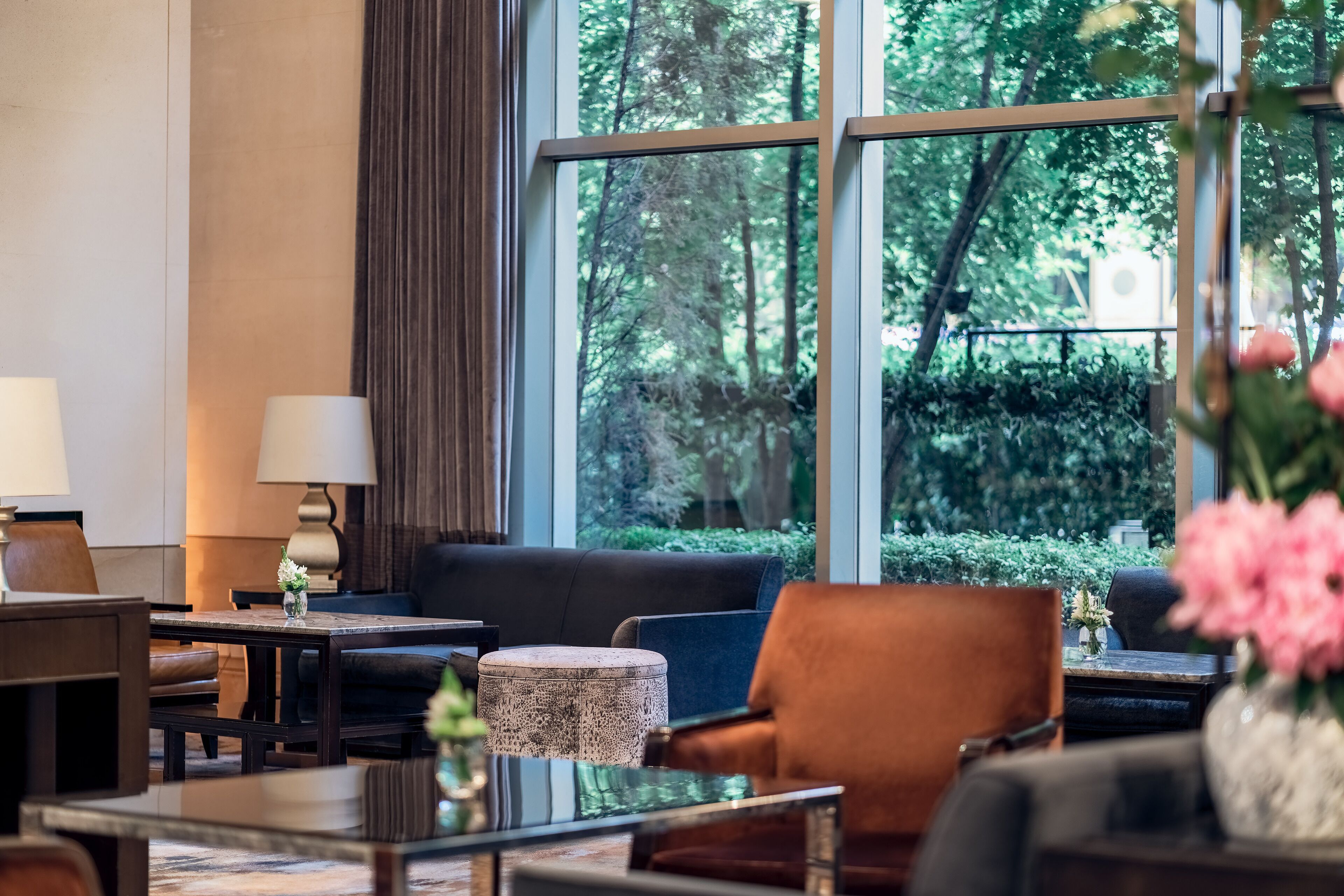 A modern hotel room with a large window offering a view of a forested area. There is a sofa, a coffee table, and a lamp on a side table.