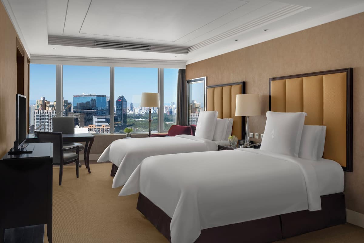 A hotel room with two beds, a desk, and a chair. There is a large window with a city view.