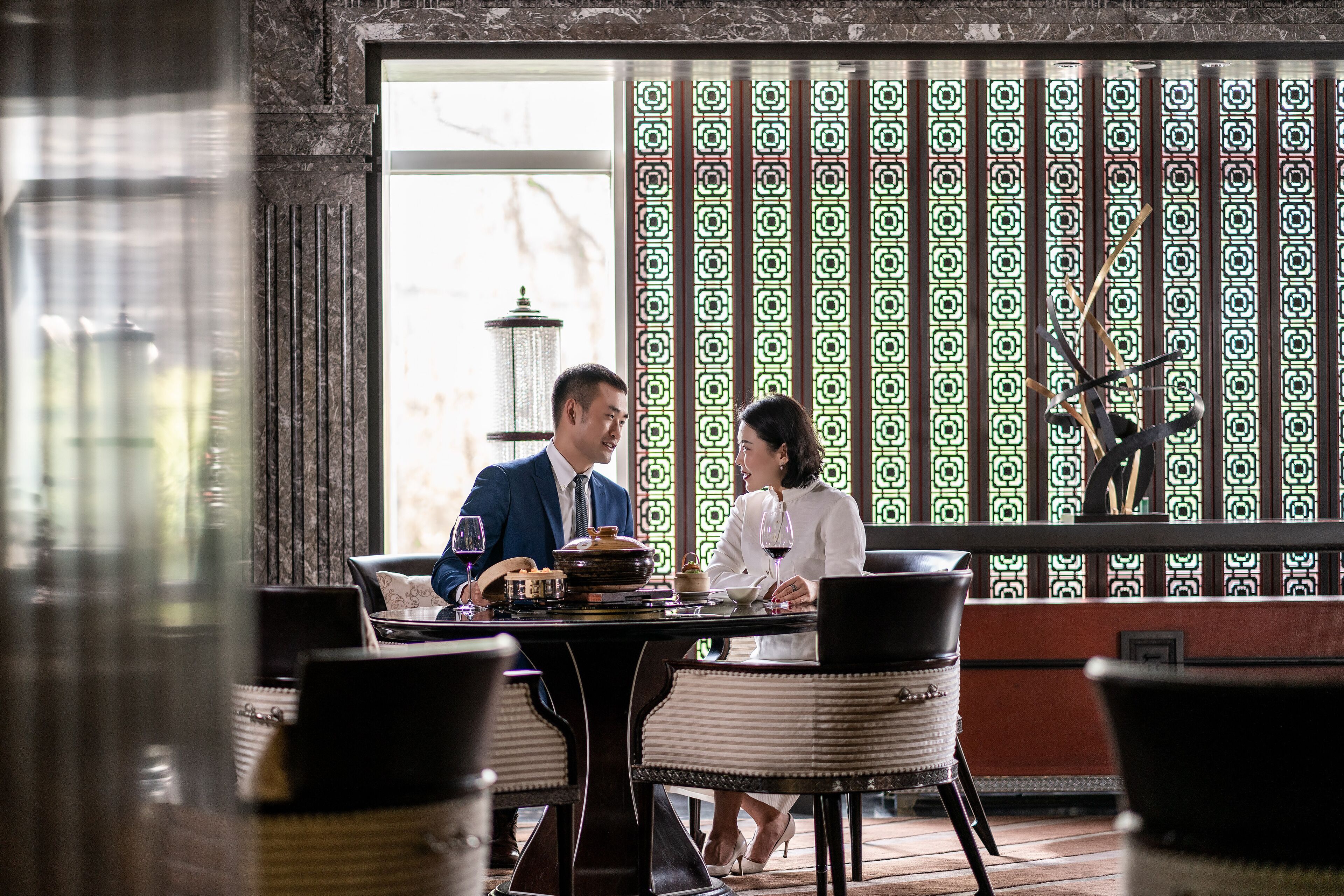 A dining area with a table set for two, a patterned wall, and a view of the outdoors.