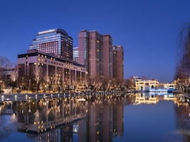 A Four Seasons Hotel with a reflection in a calm body of water, surrounded by trees and a bridge.