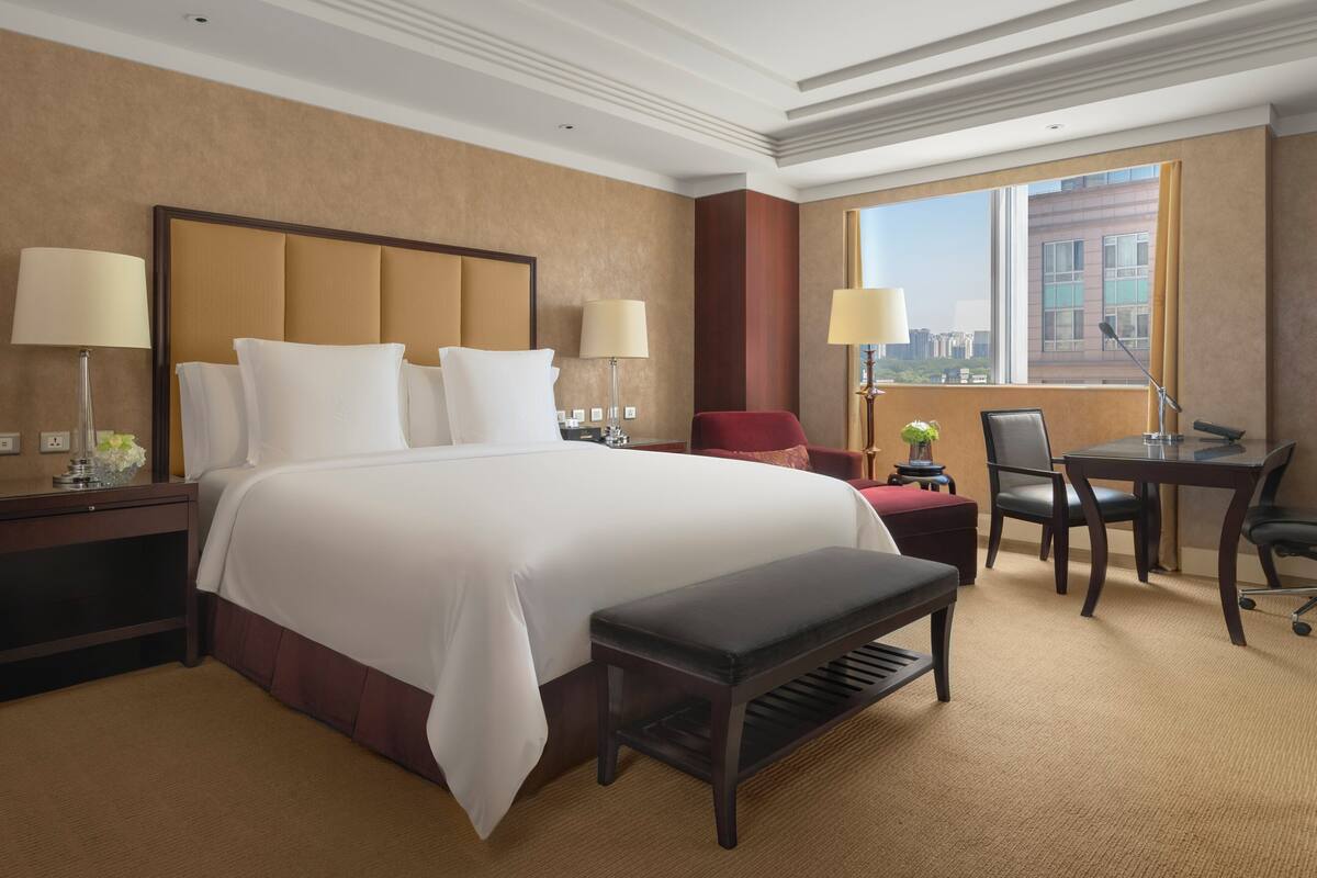 A hotel room with a large bed, two bedside tables with lamps, a desk with a chair, a red armchair, and a window with a city view.