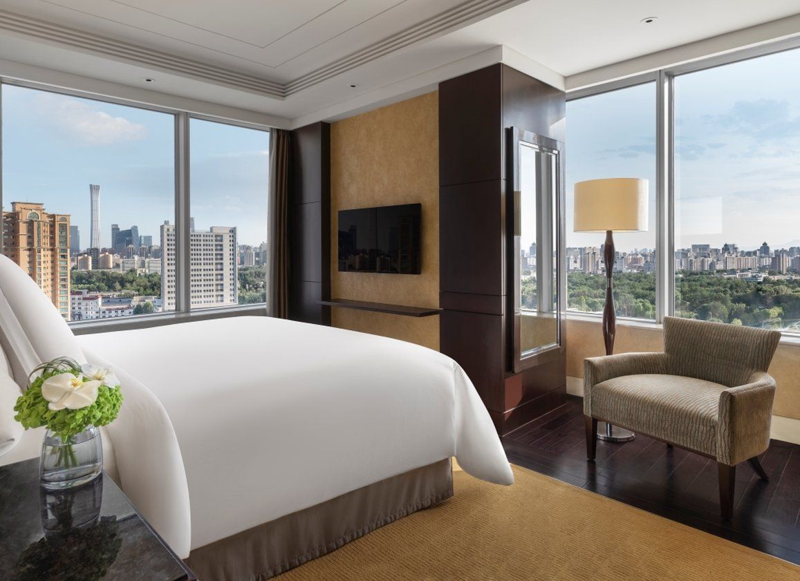 A hotel room with a large bed, a TV mounted on the wall, and a view of the cityscape through large windows.