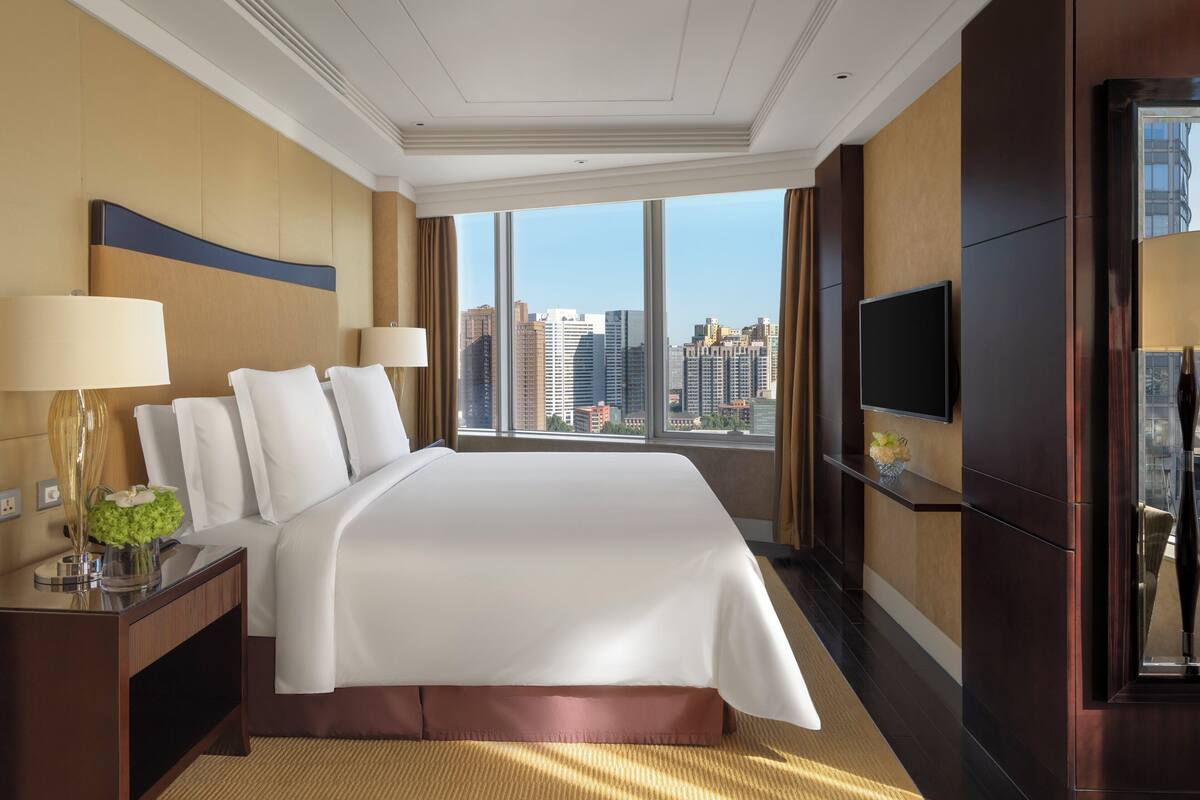 A hotel room with a large bed, a TV, a desk with a lamp, and a view of the cityscape.