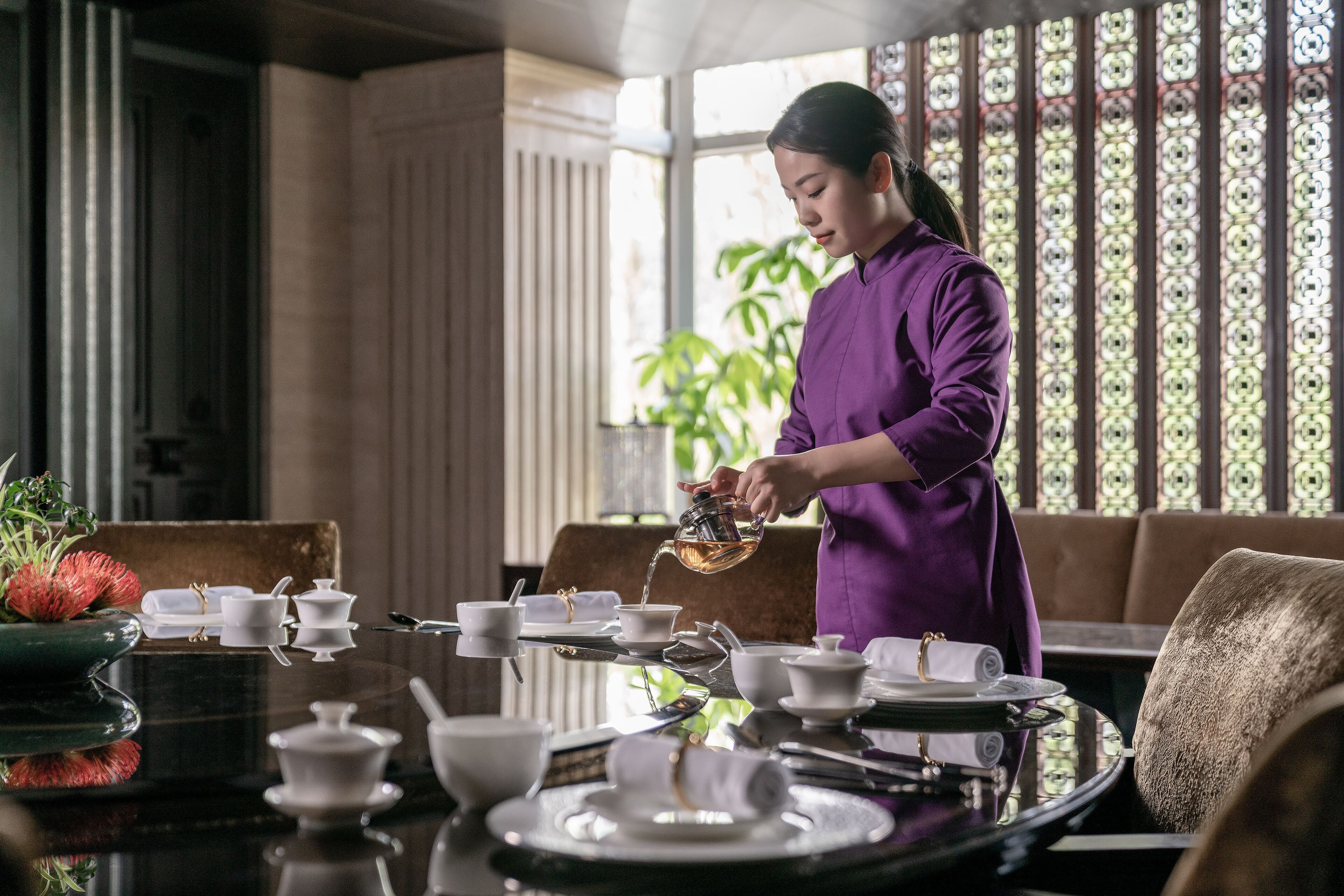 A person pouring tea into a cup at a table set with teaware and a vase of flowers.